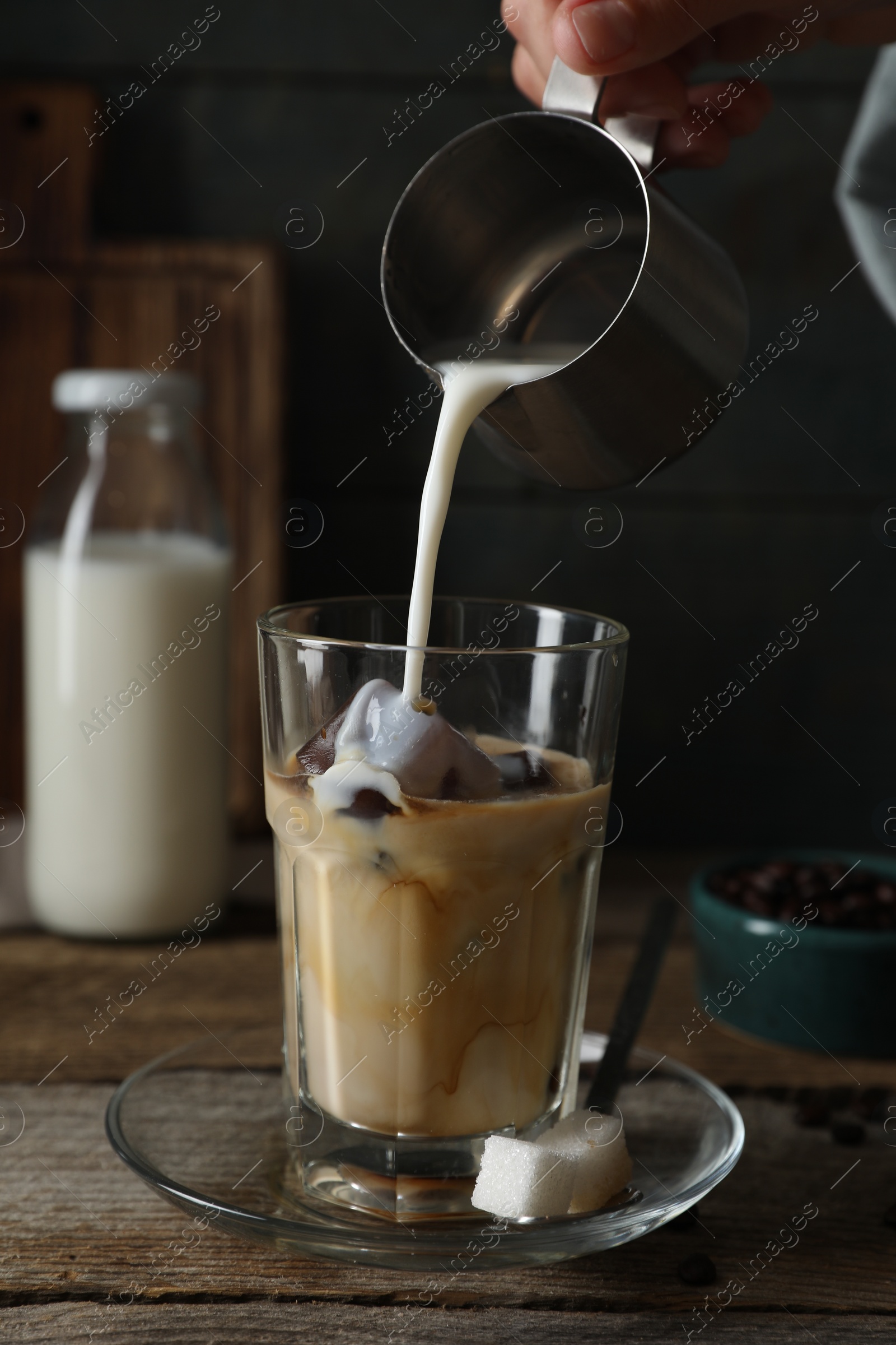 Photo of Woman pouring milk into glass with coffee ice cubes at wooden table, closeup