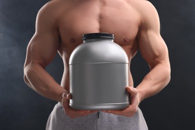 Young man with muscular body holding jar of protein powder on dark grey background, closeup