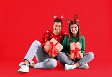 Beautiful happy couple in Christmas headbands and sweaters sitting with gifts on red background