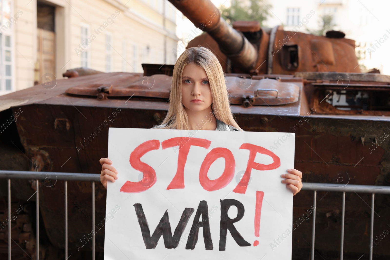 Photo of Sad woman holding poster with words Stop War near broken tank in city