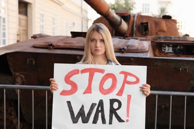 Sad woman holding poster with words Stop War near broken tank in city