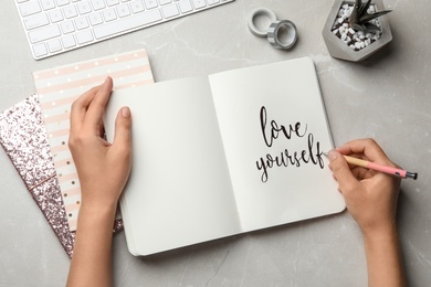 Woman writing LOVE YOURSELF in journal on grey table, flat lay