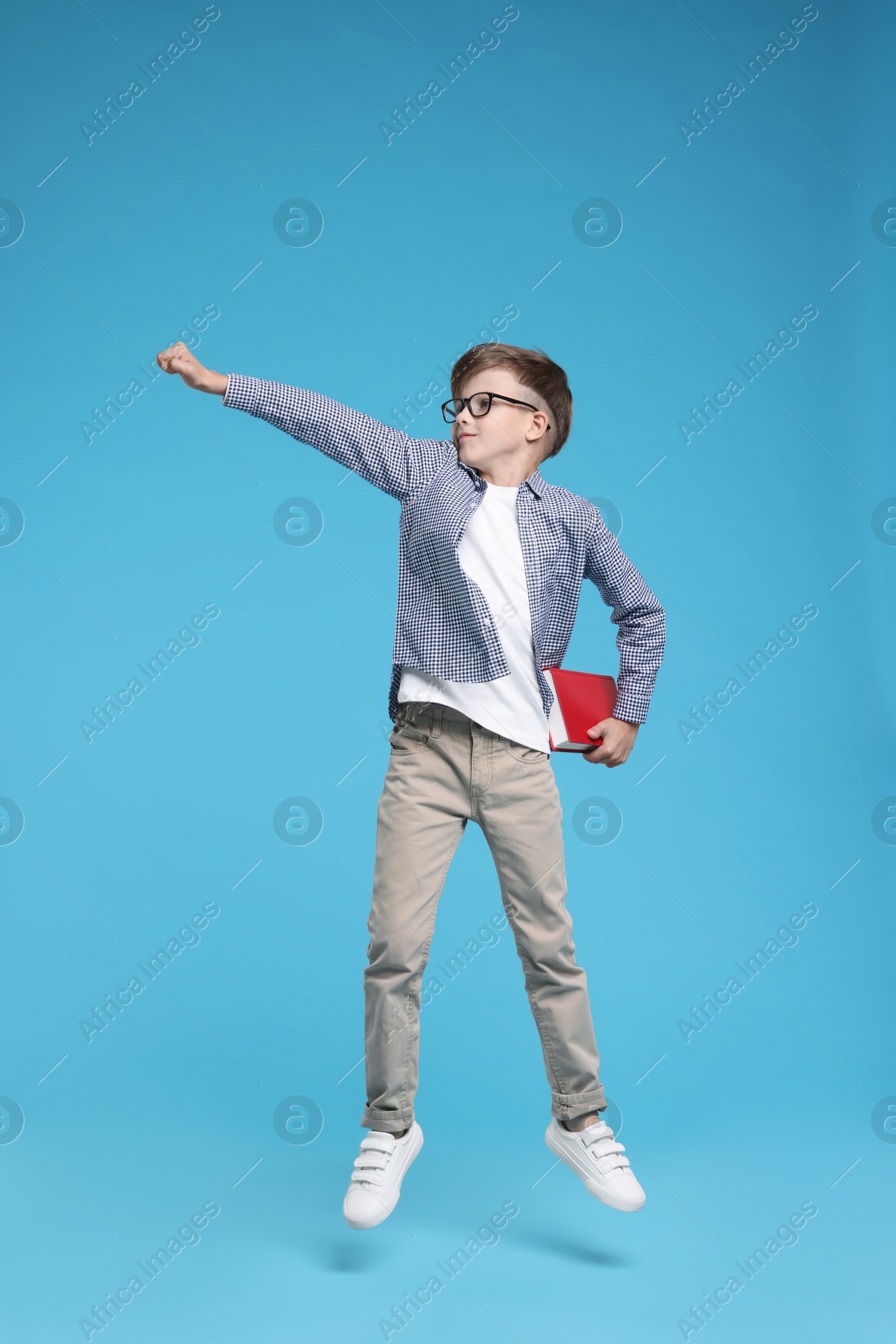 Photo of Cute schoolboy in glasses holding book and jumping on light blue background