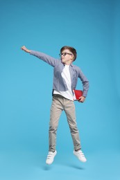 Photo of Cute schoolboy in glasses holding book and jumping on light blue background