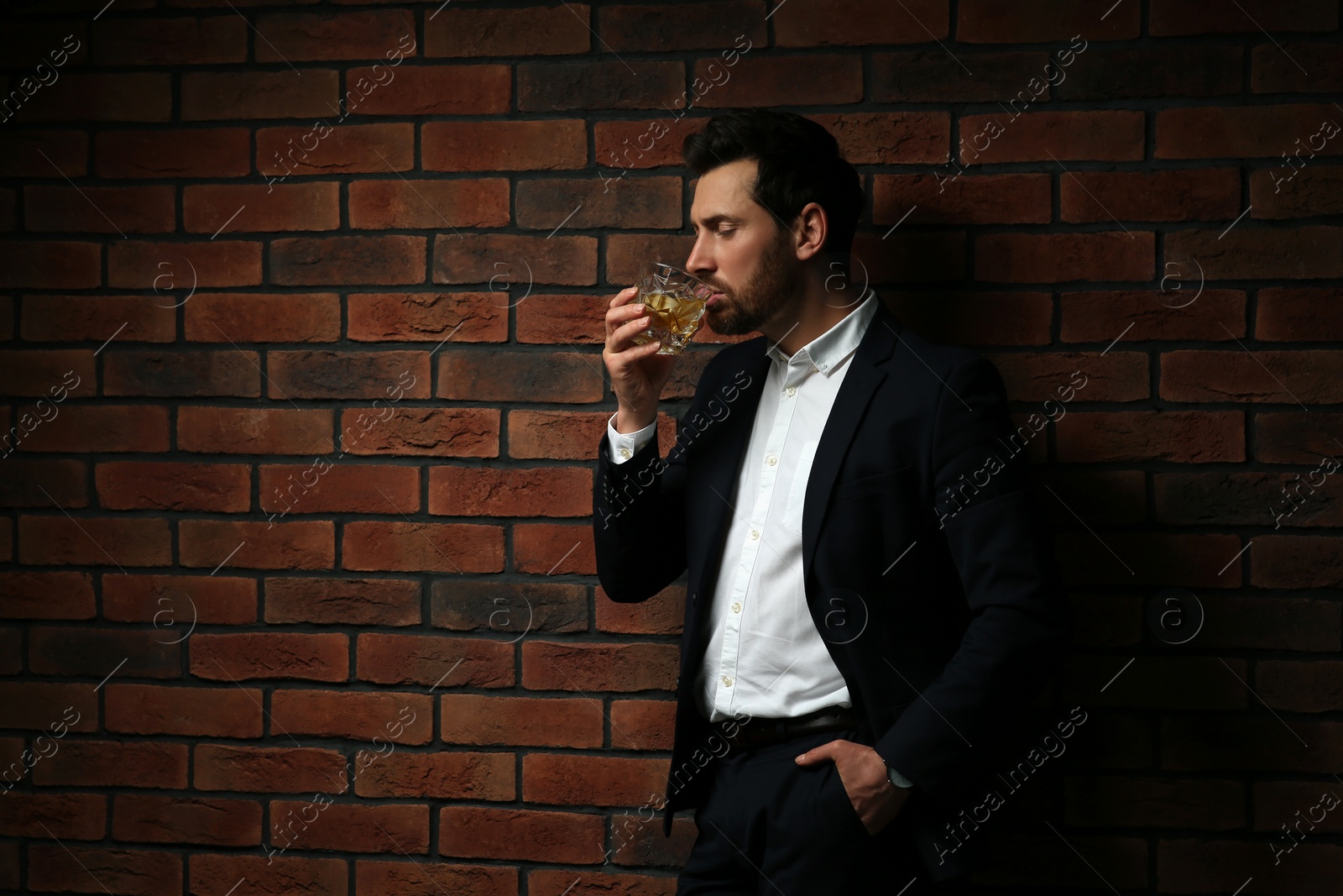 Photo of Man in formal suit drinking whiskey near red brick wall. Space for text