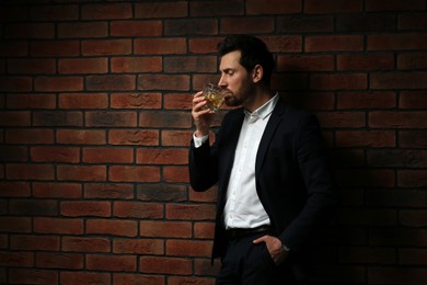 Photo of Man in formal suit drinking whiskey near red brick wall. Space for text