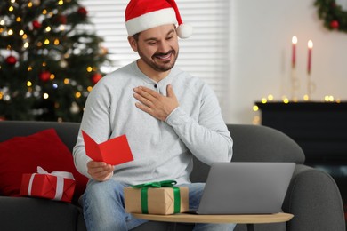 Celebrating Christmas online with exchanged by mail presents. Happy man with greeting card thanking for gift during video call on laptop at home