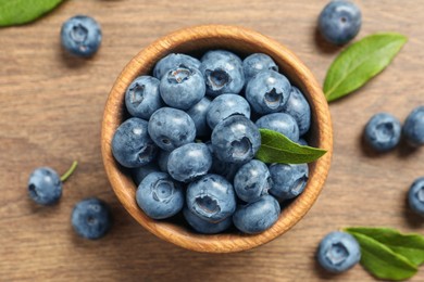 Photo of Bowl of tasty fresh blueberries with green leaves on wooden table, flat lay