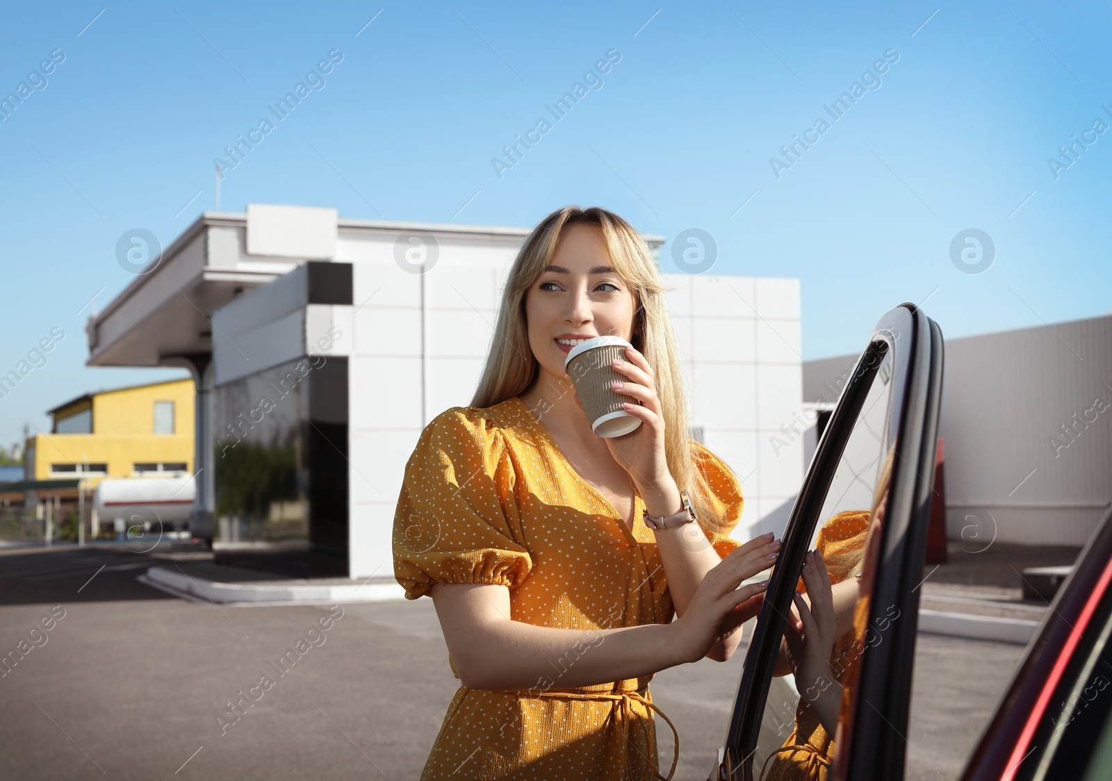 Photo of Beautiful young woman with coffee opening car door at gas station