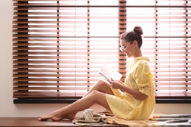 Photo of Young woman reading book near window with blinds at home. Space for text