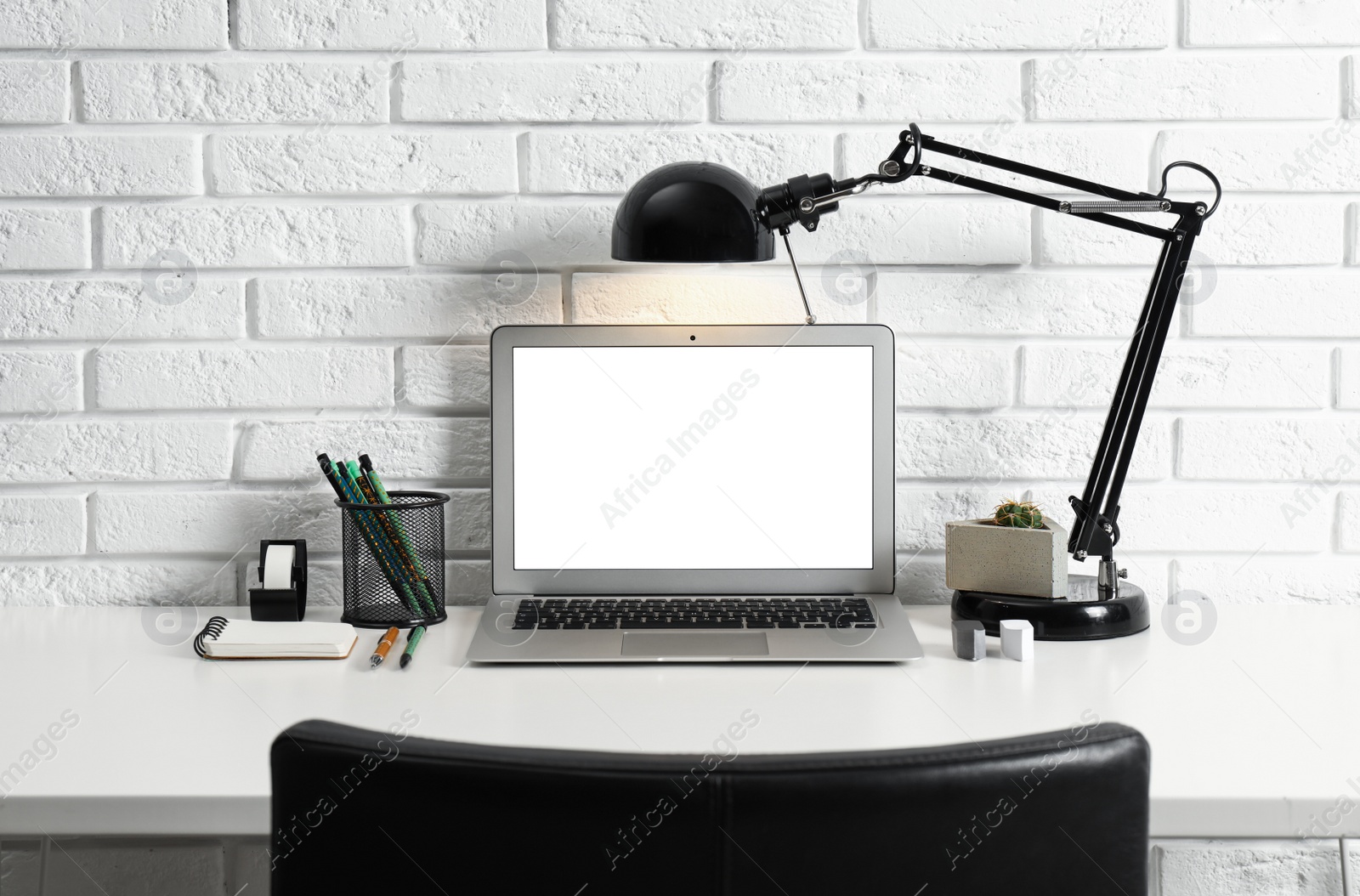 Photo of Modern workplace with laptop and lamp on table near brick wall. Space for design