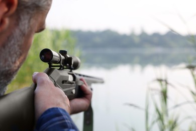 Man aiming with hunting rifle near lake outdoors, closeup. Space for text