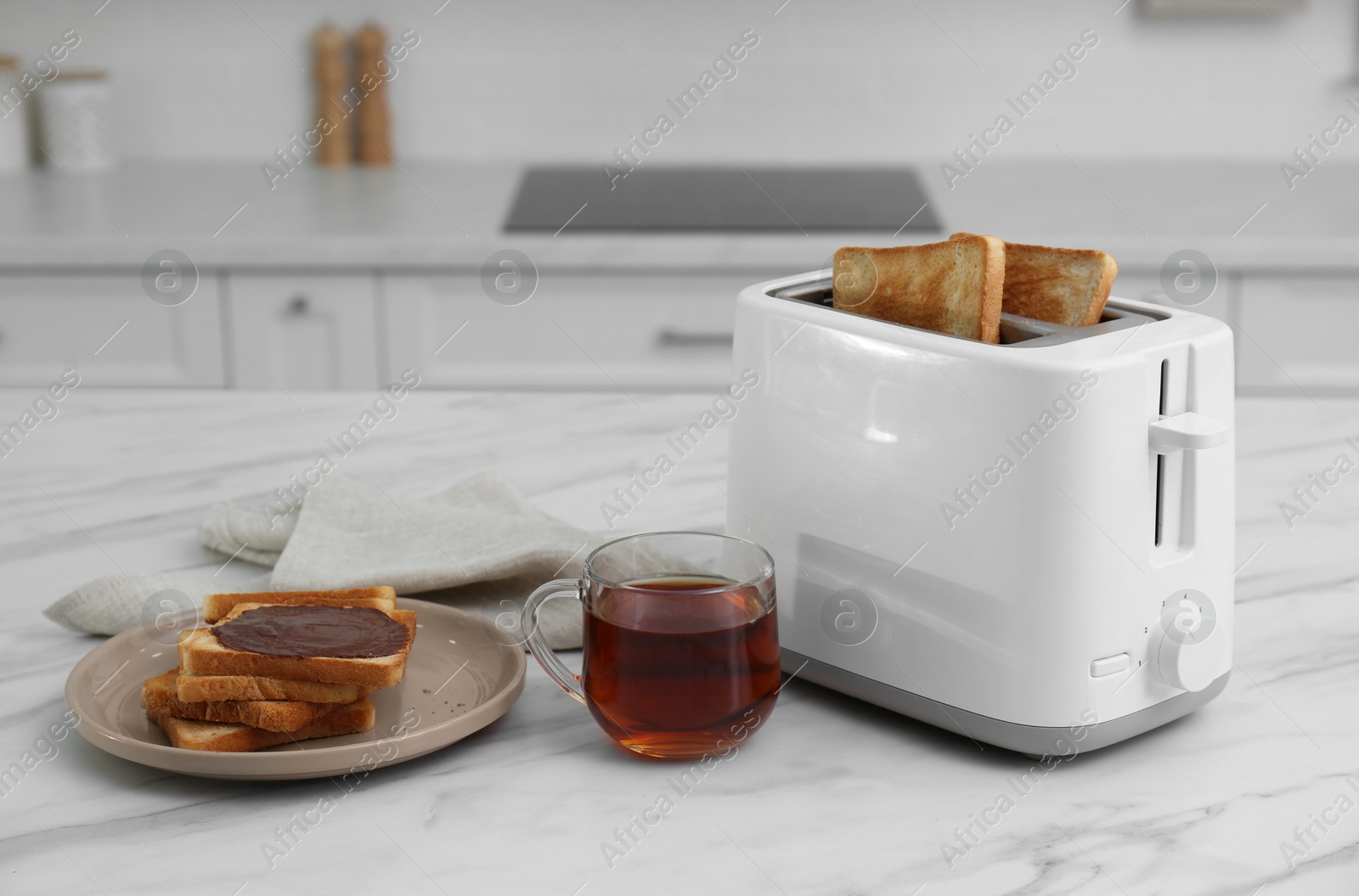 Photo of Modern toaster, bread slices with chocolate cream and cup of tea on white marble table in kitchen