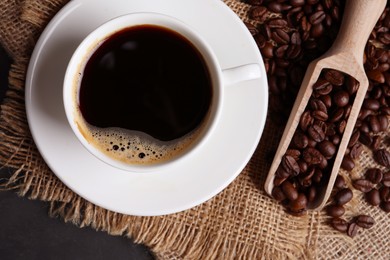 Photo of Cup of hot aromatic coffee and scoop with roasted beans on black table, top view