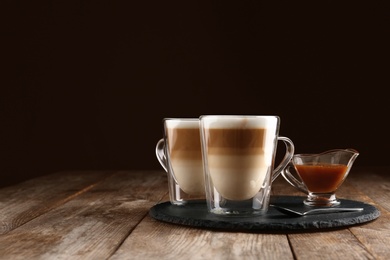 Glass cups of caramel macchiato and gravy boat with syrup served on table, space for text