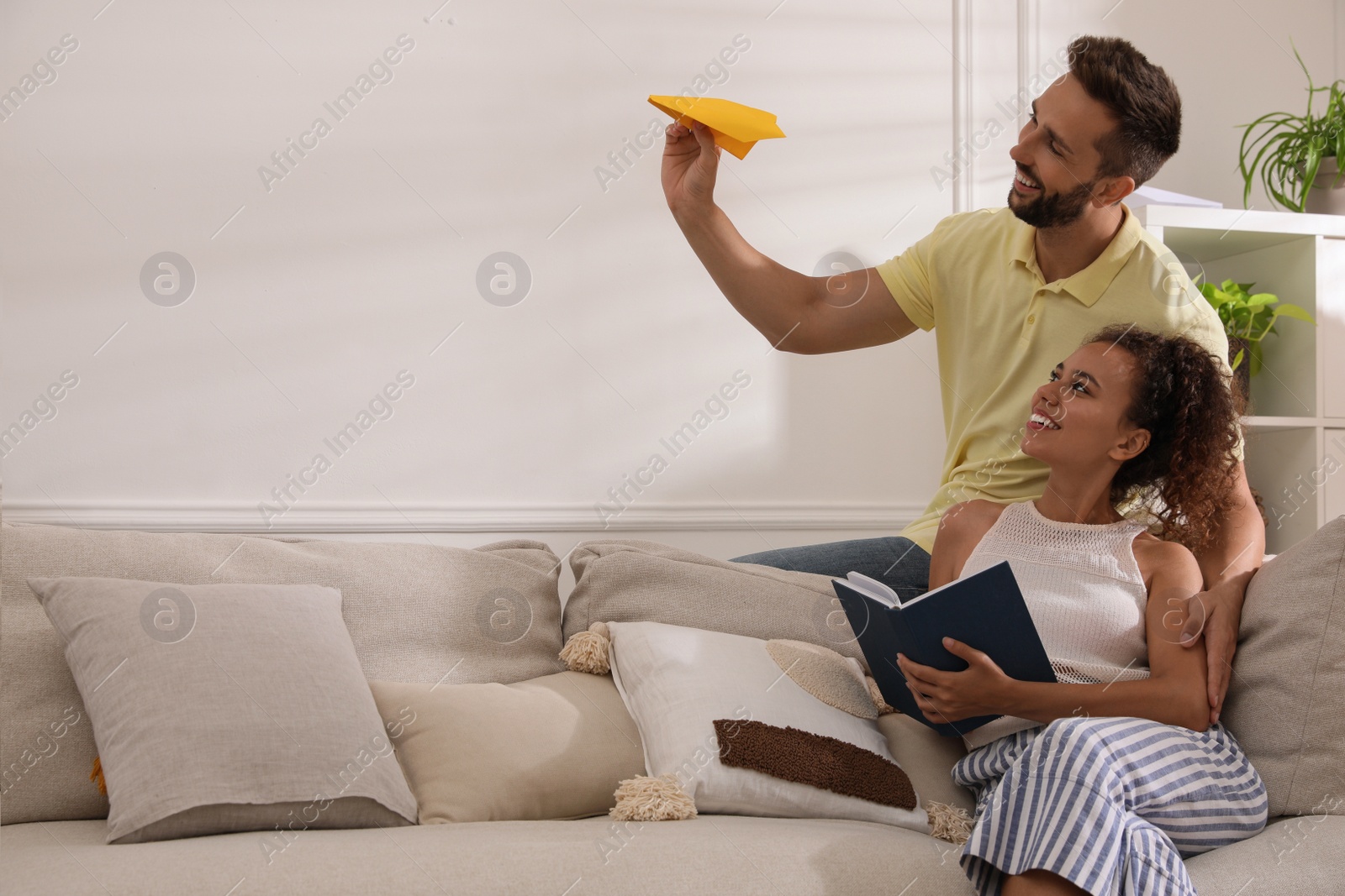 Photo of Happy man playing with paper plane while his girlfriend reading book on sofa in living room