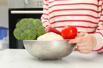 Photo of Woman holding colander with fresh vegetables at white marble table in kitchen, closeup