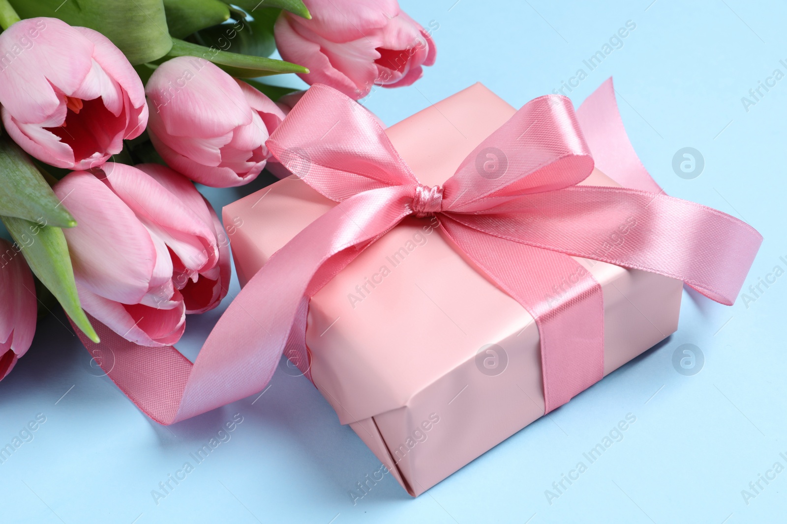 Photo of Beautiful gift box with bow and pink tulip flowers on light blue background, closeup