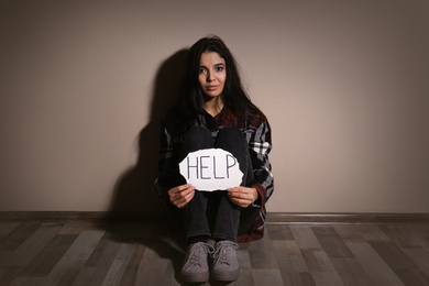Abused young woman with sign HELP near beige wall. Domestic violence concept