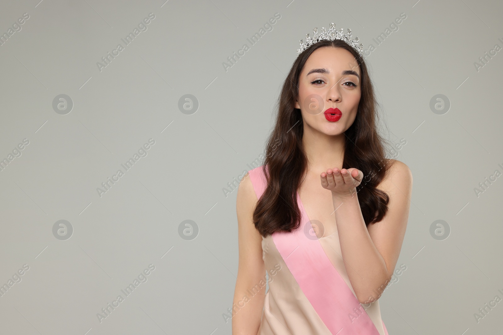 Photo of Beautiful young woman with tiara and ribbon in dress blowing kiss on grey background, space for text. Beauty contest