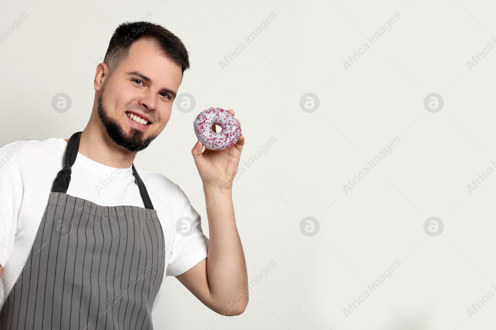 Photo of Happy professional confectioner in apron holding delicious doughnut on light grey background. Space for text