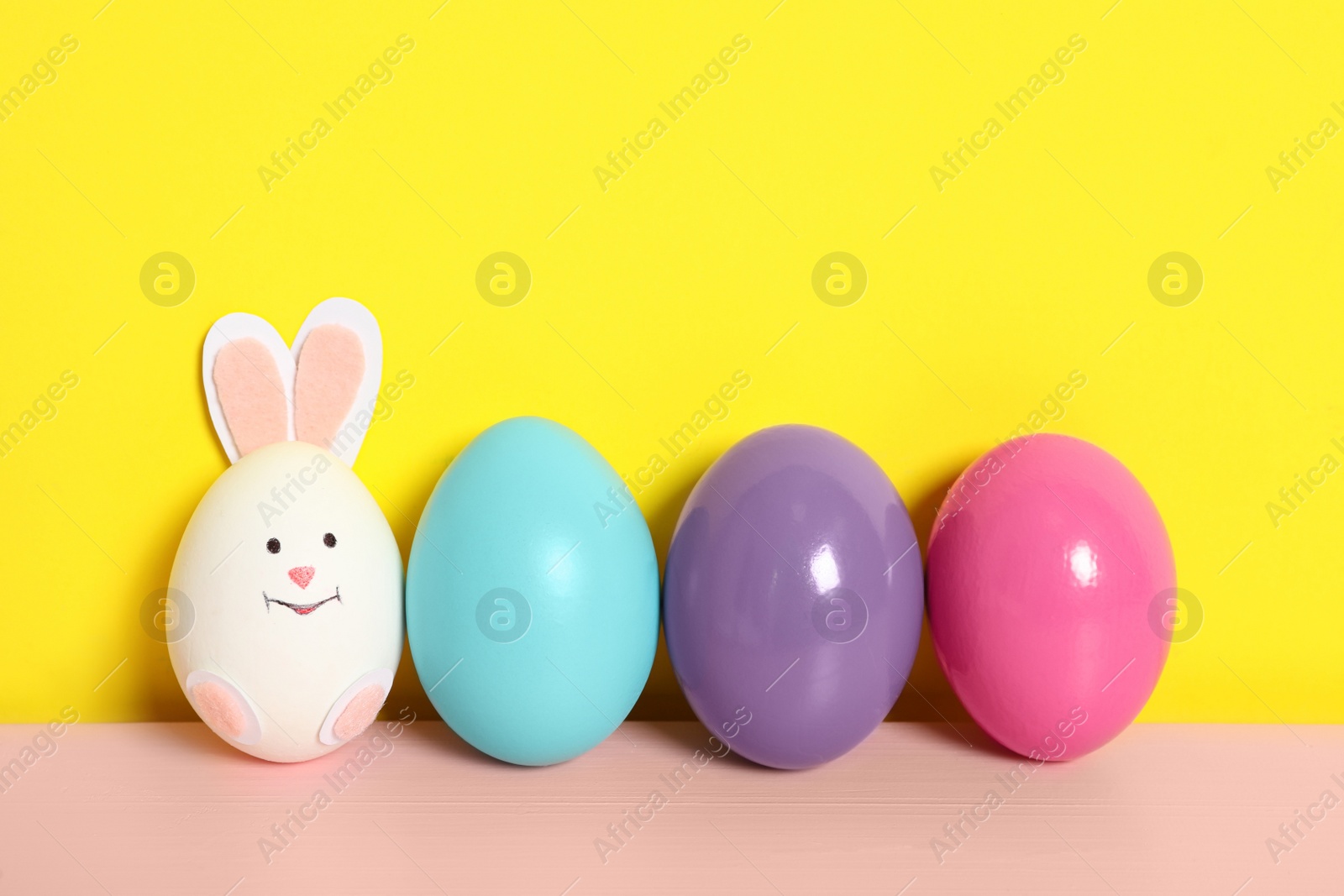 Photo of Bright eggs and white one as cute bunny on pink wooden table against yellow background, space for text. Easter celebration