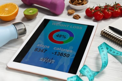 Photo of Tablet with weight loss calculator application, bottle and measuring tape on white wooden table, closeup