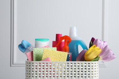 Photo of Spring cleaning. Basket with detergents, flowers and tools near white wall