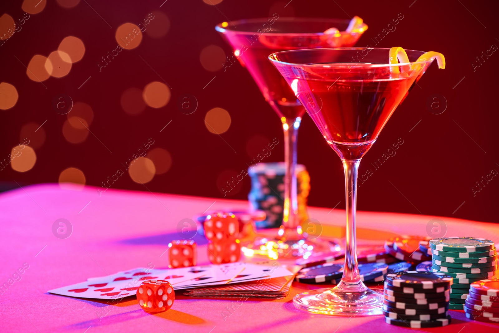 Photo of Cocktail, dice, playing cards and casino chips on table against blurred lights. Space for text