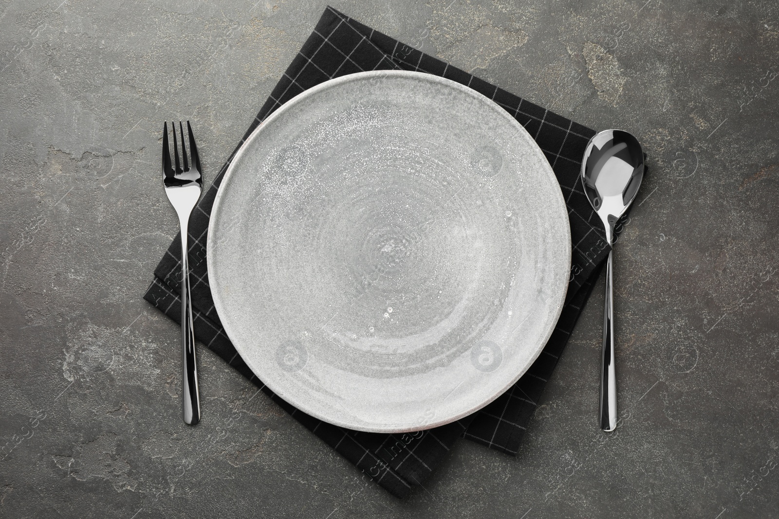 Photo of Empty plate, cutlery and checkered napkin on grey textured table, top view