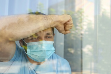 Photo of Stressed man in protective mask looking out of window, view through glass. Self-isolation during COVID-19 pandemic