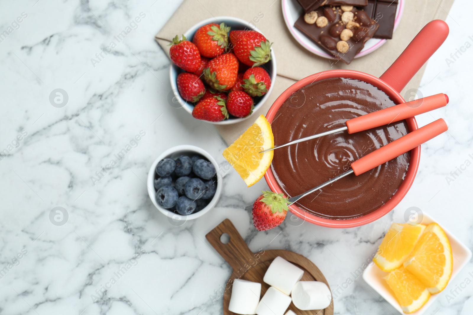 Photo of Fondue pot with melted chocolate, marshmallows, fresh orange, different berries and forks on white marble table, flat lay. Space for text