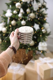 Woman with cup of delicious hot drink near Christmas tree at home, closeup