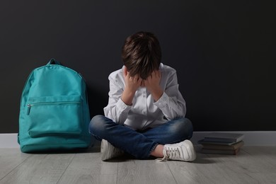 Photo of Upset boy with backpack sitting on floor near black wall. School bullying