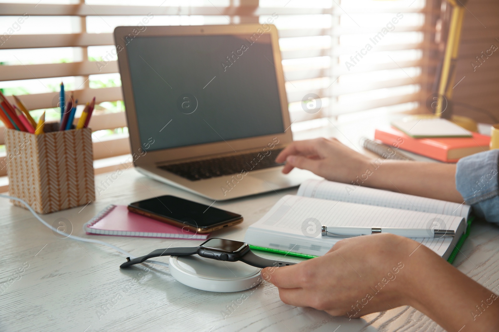 Photo of Woman putting smartwatch onto wireless charger at white wooden table, closeup. Modern workplace accessory