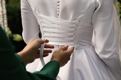 Photo of Woman helping bride to put on wedding dress outdoors, closeup