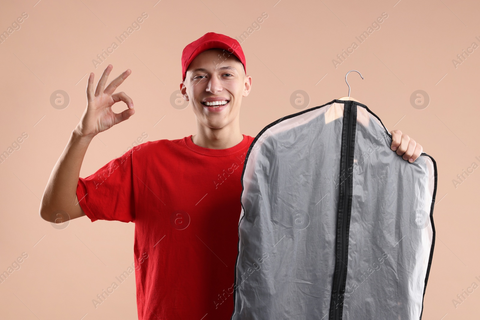 Photo of Dry-cleaning delivery. Happy courier holding garment cover with clothes and showing OK gesture on beige background