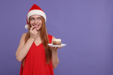 Photo of Young woman in Santa hat with piece of cake on purple background, space for text