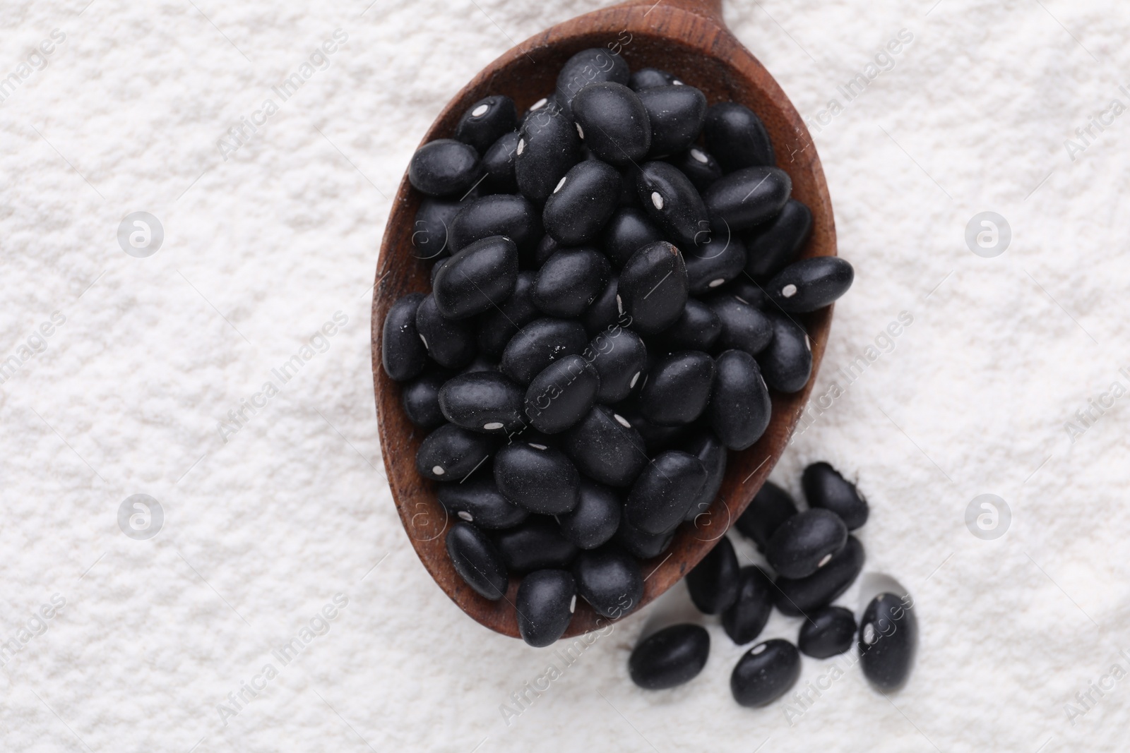 Photo of Wooden spoon with black seeds above kidney bean flour, flat lay