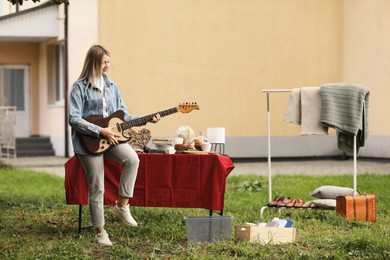 Woman holding guitar near table with different items on garage sale in yard