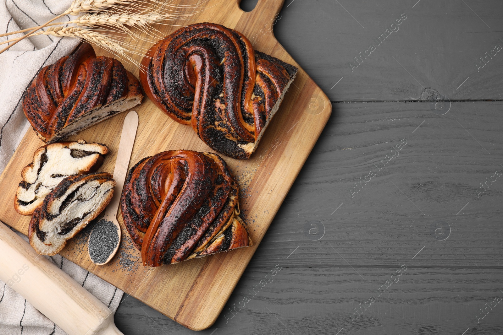 Photo of Pieces of poppy seed roll, spoon and spikelets on grey wooden table, flat lay with space for text. Tasty cake