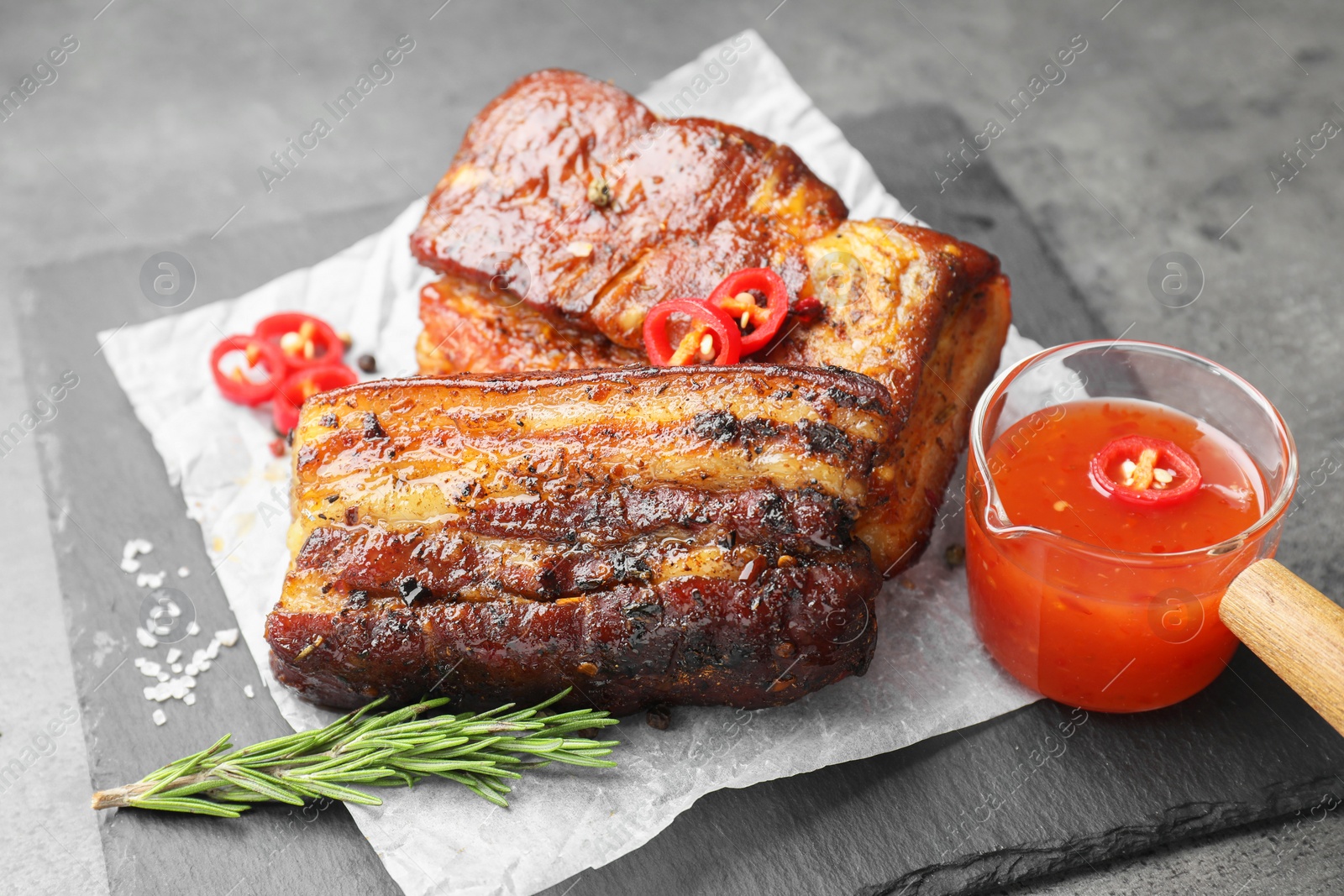 Photo of Pieces of baked pork belly served with sauce, rosemary and chili pepper on grey table