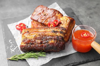 Photo of Pieces of baked pork belly served with sauce, rosemary and chili pepper on grey table