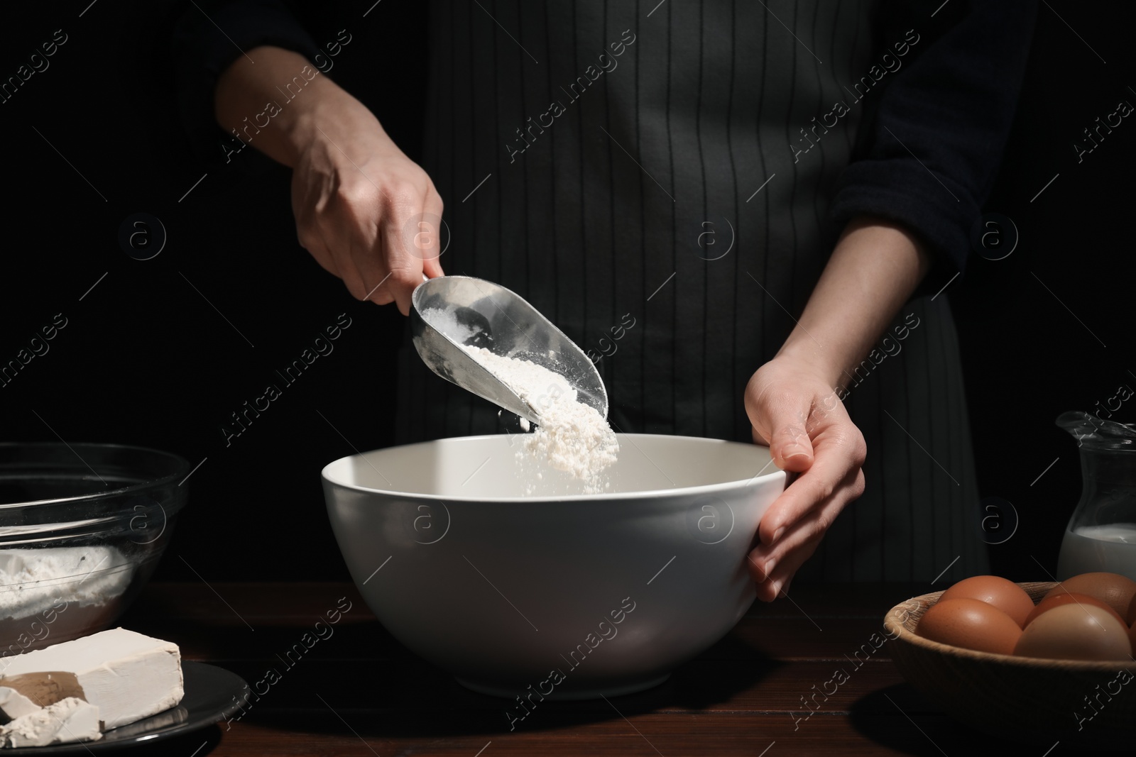 Photo of Making bread. Woman adding flour into bowl at wooden table on dark background, closeup