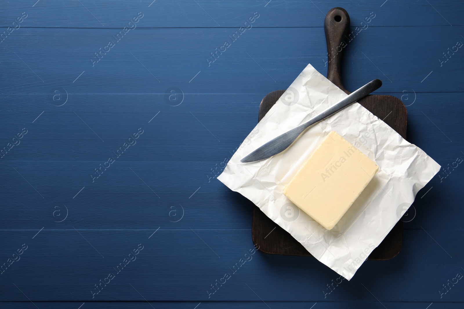 Photo of Block of tasty butter in open foil packaging and knife on blue wooden table, top view. Space for text