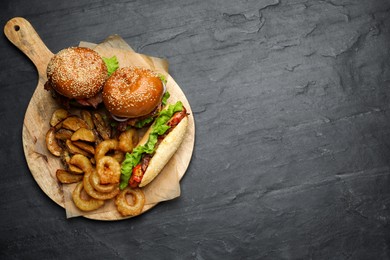 Tasty burgers, hot dog, potato wedges and fried onion rings on black table, top view with space for text. Fast food