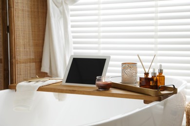 Wooden tray with tablet, cosmetic products and burning candles on bath tub in bathroom