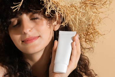 Photo of Beautiful young woman in straw hat holding tube of sun protection cream on beige background, closeup
