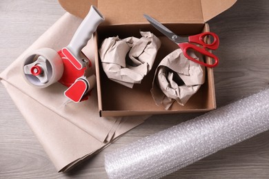 Photo of Open box with wrapped items, adhesive tape, scissors, paper and bubble wrap on wooden table, flat lay