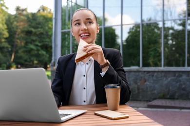 Photo of Happy businesswoman eating sandwich during lunch at wooden table outdoors